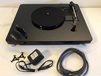 Full view of glossy black Ion ILP turntable with dock on left and accessories including power adapter, audio cable, and two 45 RPM adapters positioned in front.