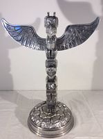 Front view of the polished metal totem pole sculpture featuring detailed eagle wings and stacked totem figures on a textured round base.
