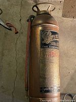 Full view of antique Pyrene soda-acid fire extinguisher showing copper body, top handle, and attached hose.