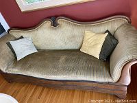 Antique wood framed sofa with beige striped upholstery, hand carved wood detail on top of the backrest, and three throw pillows (one beige, two darker). Positioned on hardwood floor against red wall with window.