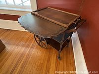 Tea trolley with one leaf folded open, showing carved wood patterns and two large rubber rim spoked wheels along with smaller support legs.