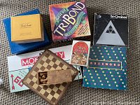 Photo showing various board games stacked and overlapping, including Monopoly, TríBond, Mille Bornes, Buzzword, Tri-Ominos, and a small wooden chess set with pieces in a storage drawer.