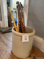 Photo showing large ceramic crock holding multiple walking canes on wooden floor near wall