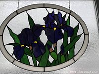 Close-up photo of an oval stained glass panel showing detailed purple iris flowers and green leaves with white and textured glass background and metal hanging chains.