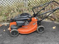 Side view of orange Ariens gas lawn mower with folded black handle, black wheels on asphalt near fence.