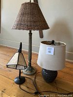 Photo of three table lamps on wooden floor. Tall lamp has woven wicker shade and twisted metal base, medium lamp has rounded dotted ceramic base with beige shade, small lamp with black metal base and geometric glass shade.