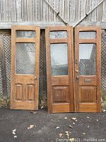 Three wooden doors with glass panels and recessed lower panels leaning against fence