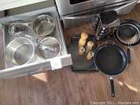 Stainless steel pots, large frying pan, glass lids, spice grinders, and metal spice rack positioned by stove and a drawer.