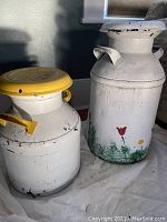 Two vintage milk cans side by side, yellow-lidded smaller can and larger white can with floral decoration