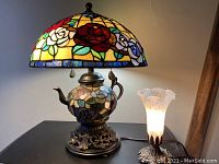 Display of two tabletop lamps: a Tiffany style teapot lamp with colorful stained glass shade and floral teapot base; and a smaller torch lamp with frosted glass shade and metal base.