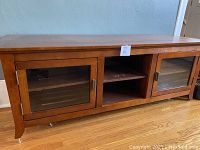 Full frontal view of the brown wood TV stand showing two glass doors and an open shelf in the center, resting on wooden floor, with inventory tag on top center