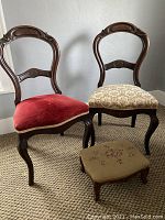 Two Victorian wooden chairs, one with red velvet upholstery and the other with beige floral fabric. A small footstool with floral needlepoint upholstery is in front of the chairs.