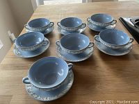 Seven sets of blue Wedgwood jasperware soup bowls each paired with matching saucers, arranged on a wooden surface.