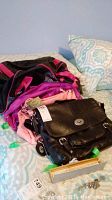 Photo of four bags laid on a bed, including a black Fossil satchel, pink Isabelle handbag, purple Nike backpack, and black/pink Roots backpack.