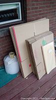 Stack of flat cardboard boxes of various sizes against brick wall on porch floor with bubble wrap roll and tape gun and packing tape on floor beside them.
