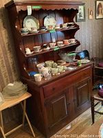 Front view of vintage wooden cabinet showing scalloped backboard, two plate rail shelves with numerous decorative plates and teacups, and lower cabinet doors.
