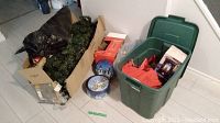 Showing the 6ft pre-lit Christmas tree parts, green plastic storage bin filled with Christmas decorations including wrapping paper, boxed items, and loose ornaments on the floor.