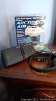 Image of Arctic Air Ultra-Pro air cooler box, black digital clock radio, and black headphones with cable on wooden surface.