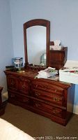 Frontal view of the dark wood dresser with several drawers and an arch-top mirror attached. Several personal items and clutter on top of dresser.
