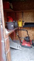 Photo of red Canadian Tire gas lawn mower with handle, plastic watering cans, plant pot, jerry cans, and some miscellaneous items in a garden shed