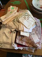 Top-down view of a varied stack of table linens, napkins, placemats, and cloths, showing embroidery, lace, and patterned designs in creams and beiges.