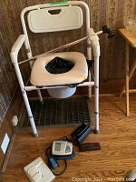 White commode chair with padded seat, armrests and removable bucket, with items on floor nearby