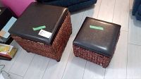 Two wicker ottomans of different sizes, both with dark brown faux leather lift-up storage tops, displayed on light flooring near couch.