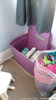 Purple plastic bin placed in corner with open lid, containing puzzles and board games including Cranium and Crayola.