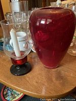 Photo showing large red Christmas vase and glass hurricane candle holder with black base and white wax candle inside, on wooden table