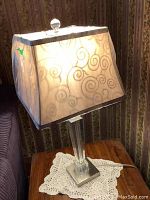 Photo showing one illuminated crystal base lamp with square beige fabric shade featuring swirl patterns and a round clear finial on top.