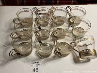 Set of 12 clear glass Greek teacups each with a metal holder with Greek key gold design, plus one extra metal holder, arranged on table.