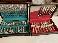 Two open wooden cases containing incomplete sets of silver plated silverware. One case has a blue interior and twisted handle pattern set; the other case has a red interior and plain handle set.