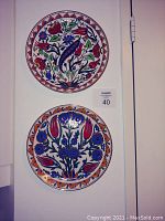 Two ceramic decorative wall plates shown hanging on a wall, one above the other, both with colorful hand painted floral and peacock designs.