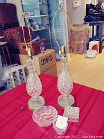 Two cut glass table lamps and three cut glass decorative items on red tablecloth. Lamps are teardrop shaped with diamond cut glass bases and brass fittings. Small bowls and small box with lid also visible front and center.