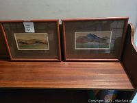 Two original watercolour mountain landscape paintings in wood frames with glass and mats, set on wooden bench.
