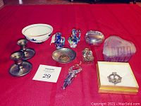 All items arranged on red cloth including candle holders, ceramic birds, metal dish, lidded box, pink glasses, bottle stoppers, bowl and brooch.