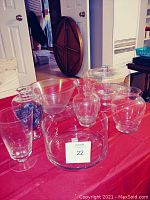 Nine clear glass items on red tablecloth including a glass pedestal cake stand, vases, bowls, and decorative glass piece with blue accents