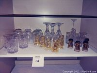 Full view of shelves with assorted stemware, liquor shot glasses in amber tint, clear crystal rock glasses, two large glass decanters with stoppers.