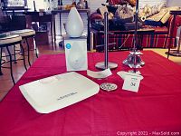 7 bathroom accessory items on red cloth including essential oil diffuser, handcloth tree, toilet roll stand, soap dishes, compact mirror, and scale