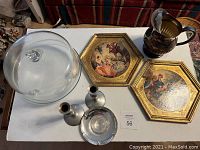 Photo showing six items on a white surface: glass cake stand with dome lid, silver-plated jug, pair of silver-colored candle stick holders, two hexagonal framed art pieces with gold-tone borders and painted scenes.
