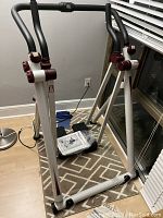 Health Walker exercise machine showing dual handlebars, white and maroon metal frame, and stable base used for fitness or rehabilitation.