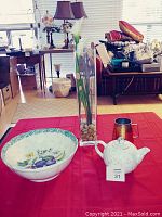 Photo showing large ceramic bowl, porcelain teapot labeled with number 31, rectangular glass vase with pebbles and flowers, and copper stein on a red cloth background with room interior visible