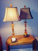 Pair of acrylic table lamps on a wooden table against a blue wall; lamps have multi-colored acrylic bases and beige fabric shades