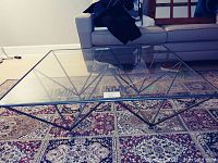 Full view of square glass top coffee table with black geometric wrought iron legs on a patterned carpet in front of a sofa.