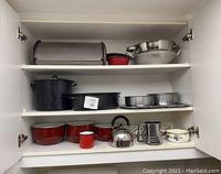Front view of open white kitchen cabinet with three shelves showing variety of metal kitchen bowls, pots, pans, kettle, and baking pans all stacked neatly inside.