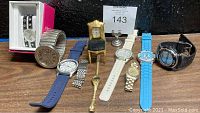 Photo of assortment of nine watches arranged in a row on a wooden surface including a decorative miniature chair clock