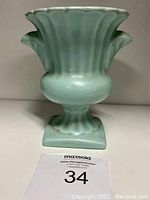 Front view of turquoise ceramic vase with flared top and pedestal base, with auction number card '34' in front