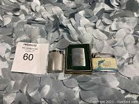 Three vintage lighters shown on silver fabric, includes textured silver lighter, boxed Columbia Production Company lighter, and Blue Dolphin advertising lighter