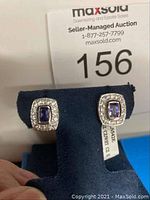 Pair of earrings displayed on blue velvet stand with auction number 156 in background. Earrings feature rectangular blue stones surrounded by small white stones.