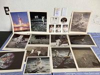 Overview photo showing about 15 large NASA prints including moon walk scenes, lunar module, Saturn V rocket launch, Earth from space, and astronaut portraits plus commemorative badges